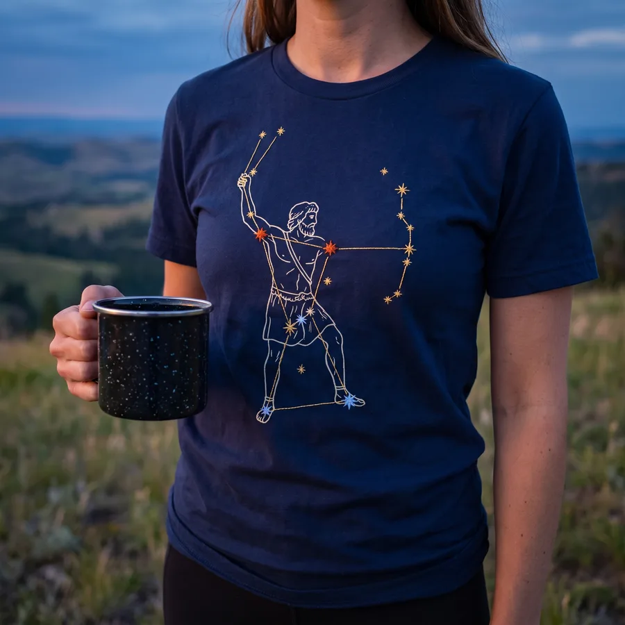 A customer on a hillside at dusk wearing the navy Orion the Hunter tee, holding an enamel mug